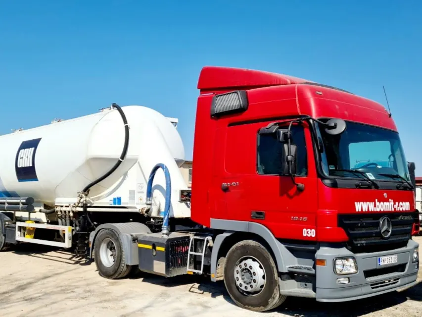 Crveni tegljač Mercedes-Benz Actros sa belom cisternom za cement, na zemljanom platou pod vedrim nebom.