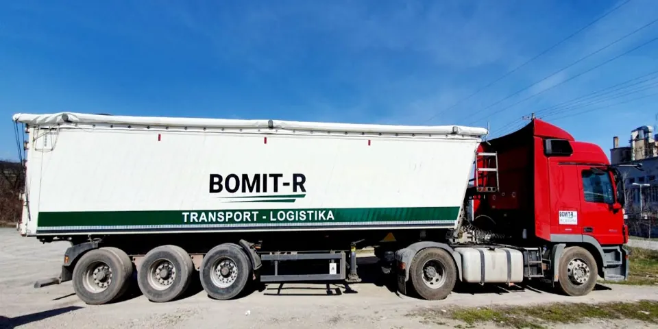 Crveni tegljač sa belom poluprikolicom kiperom i natpisom BOMIT-R Transport – Logistika, na platou pod vedrim nebom.