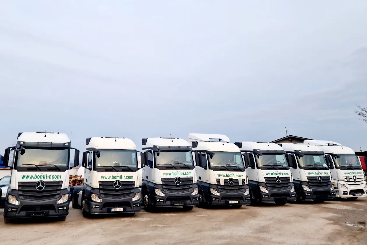 Sedam belih tegljača Mercedes-Benz Actros sa natpisom www.bomit-r.com, poređanih u nizu na šljunkovitom platou pod oblačnim nebom.