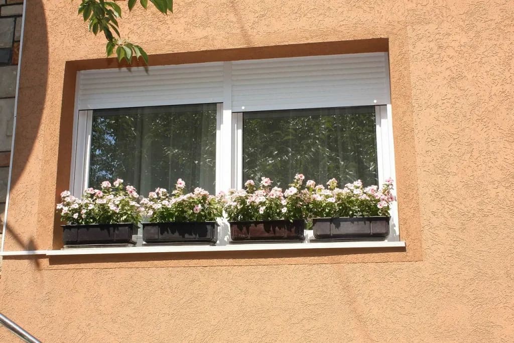 Dvokrilni beli PVC prozor sa spoljnim roletnama i žardinjerama sa cvećem na svetloj fasadi.