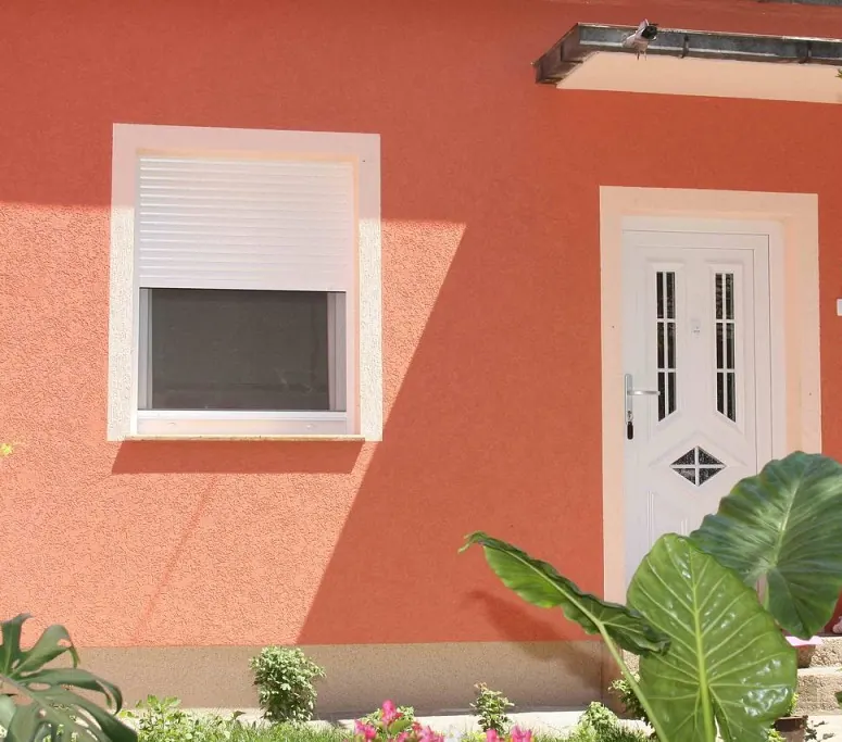 Bela PVC ulazna vrata sa ukrasnim panelom i prozor sa spoljnom roletnom na fasadi boje terakote.