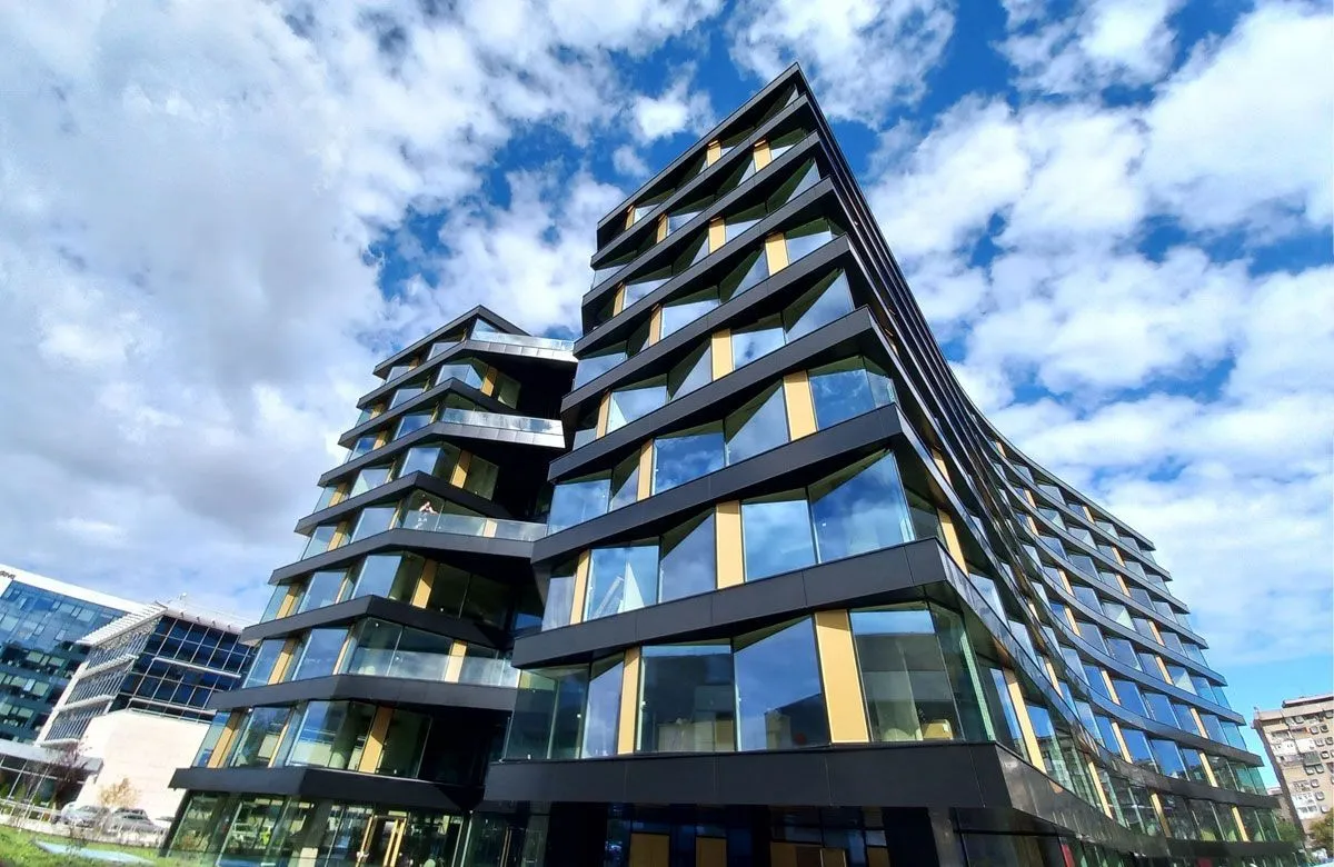 Poslovna zgrada GTC X na Novom Beogradu, stepenasto uvučenih staklenih fasada sa crnim okvirima, fotografisana odozdo pod oblačnim nebom.