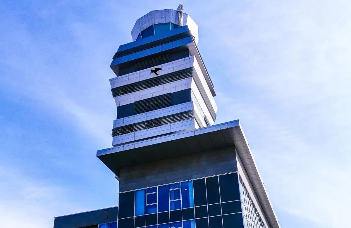 Toranj kontrole letenja beogradskog aerodroma, bela kula sa staklenom kabinom na vrhu, snimljena odozdo pod vedrim nebom.
