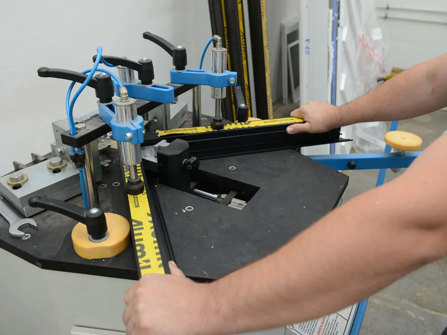 Ruke radnika koji na mašini za spajanje uglova sklapa ram od aluminijumskog profila sa žutom zaštitnom folijom.