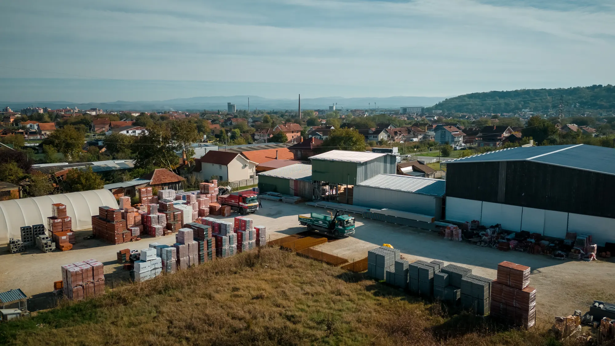 Stovarište Promax Trade u Jagodini iz vazduha: palete zidnih blokova, magacinske hale i kamion za dostavu, a u pozadini grad i brda.