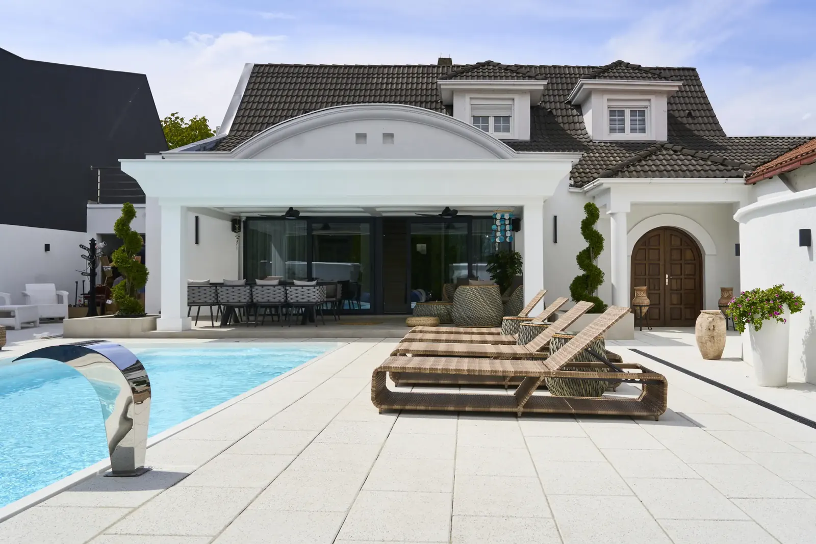Terasa uz bazen popločana velikim svetlim betonskim pločama Linia Urbana, sa ležaljkama ispred bele kuće.